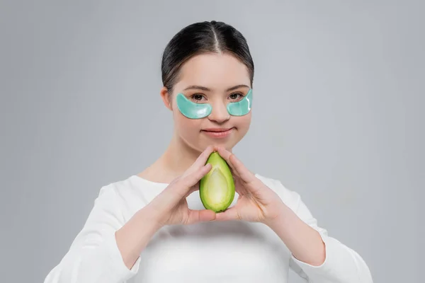 Mujer sonriente con síndrome de Down y parches oculares con aguacate aislado en gris - foto de stock