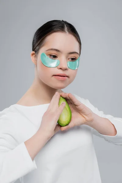 Woman with down syndrome and eye patches holding avocado isolated on grey — Stock Photo