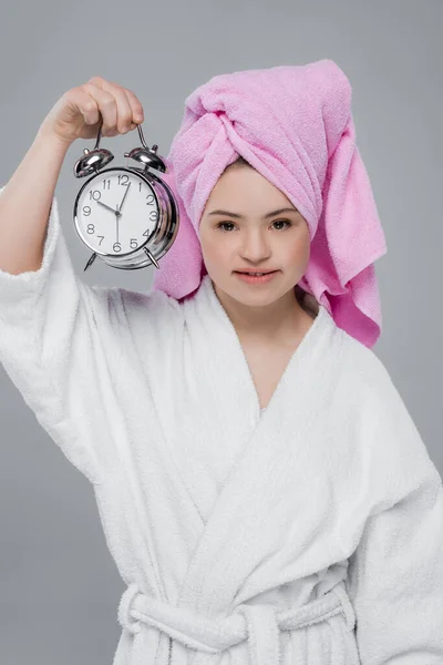 Modelo sonriente con síndrome de Down en albornoz con reloj despertador aislado en gris - foto de stock