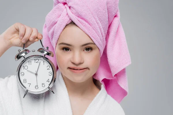 Woman with down syndrome in bathrobe and towel holding alarm clock isolated on grey — Stock Photo