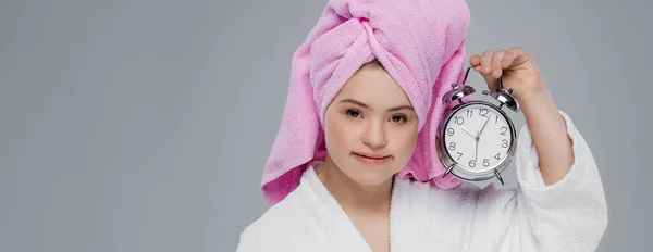 Junge Frau mit Down-Syndrom im Bademantel und Handtuch auf dem Kopf, Wecker isoliert auf grau, Banner — Stockfoto
