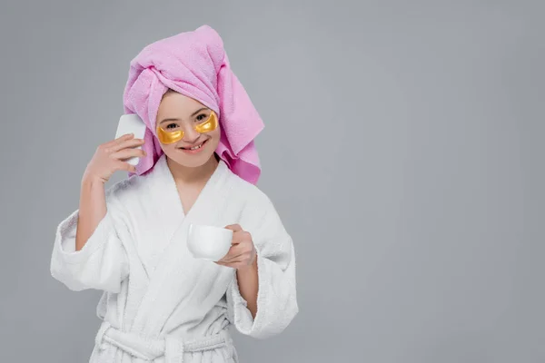 Femme souriante avec syndrome du duvet en peignoir et cache-yeux tenant du café et parlant sur un téléphone cellulaire isolé sur gris — Photo de stock