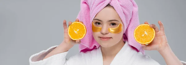 Молодая женщина с синдромом Дауна в халате и глазных повязках держит оранжевый изолирован на сером, баннер — стоковое фото