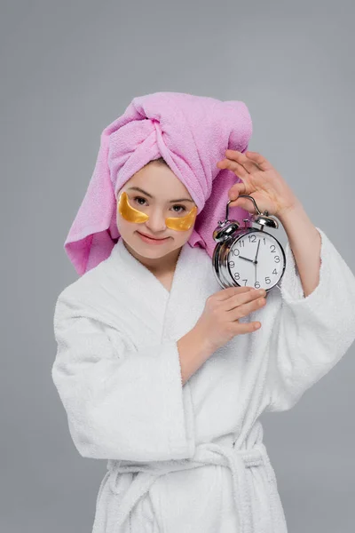 Woman with down syndrome in eye patches and bathrobe holding alarm clock isolated on grey — Stock Photo