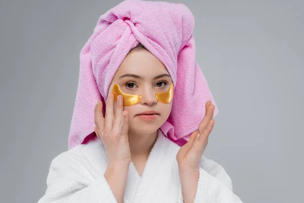 Woman with down syndrome in towel and bathrobe applying eye patches isolated on grey — Stock Photo