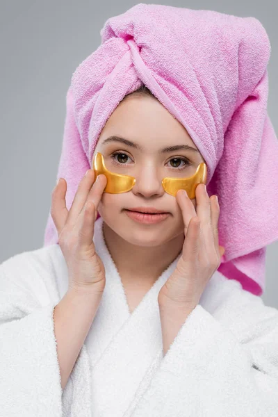 Retrato de mujer joven con síndrome de Down en albornoz y toalla aplicando parches oculares aislados en gris - foto de stock
