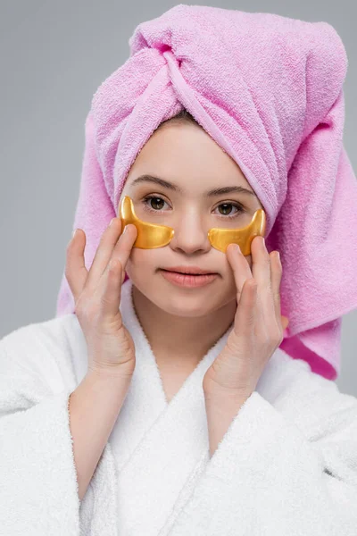 Portrait of young woman with down syndrome in bathrobe and towel applying eye patches isolated on grey — Stock Photo