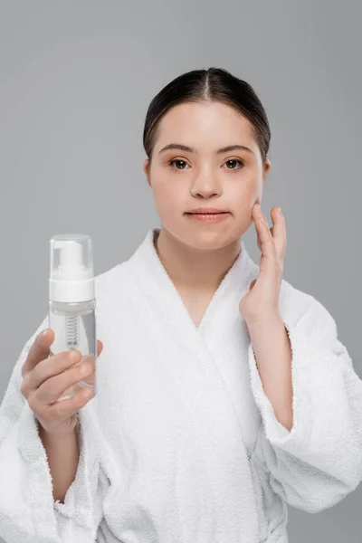 Young woman with down syndrome in bathrobe holding cleaning foam and touching face isolated on grey — Stock Photo