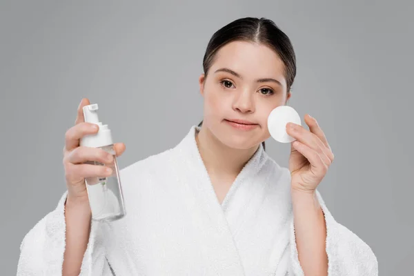 Woman with down syndrome in bathrobe holding cleansing foam and cotton pad isolated on grey — Stock Photo