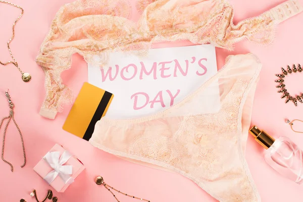 Top view of card with womens day lettering near credit card, lingerie and perfume on pink background — Stock Photo