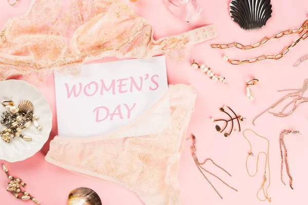Top view of card with womens day lettering near golden accessories and lingerie on pink background — Stock Photo