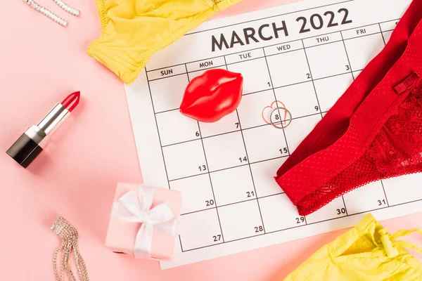 Vista superior de lencería cerca del presente, calendario de marzo y lápiz labial sobre fondo rosa - foto de stock