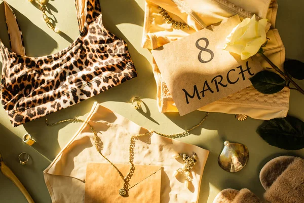 Vue du dessus de lingerie, rose près des pantoufles et carte avec lettrage 8 mars sur fond vert — Photo de stock