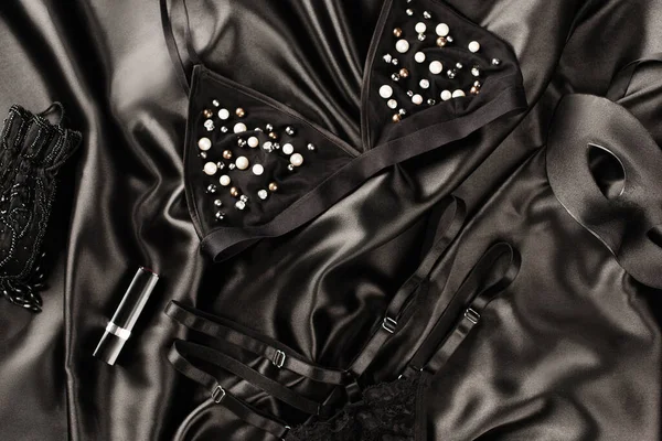 Vue du dessus de lingerie sexy près du masque et du rouge à lèvres sur fond de satin noir — Photo de stock