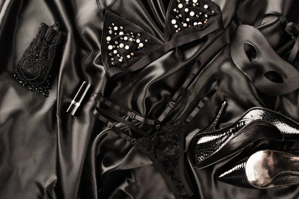 Вид сверху на сексуальное белье, маску и каблуки на черном сатиновом фоне — стоковое фото