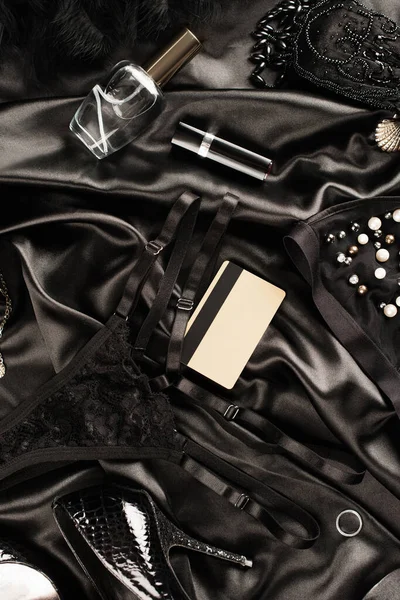Vista superior de la tarjeta de crédito cerca de bragas sexy, perfume y talón sobre fondo satinado negro - foto de stock