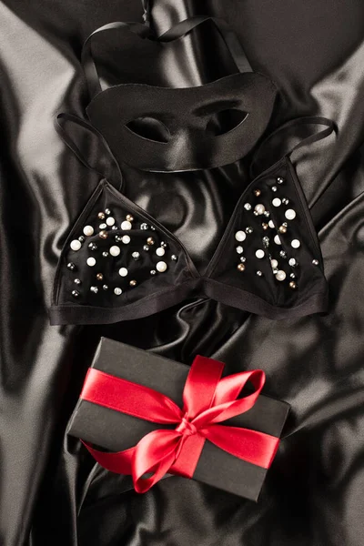 Visão superior do presente perto de sutiã e máscara sexual no fundo de cetim preto — Fotografia de Stock