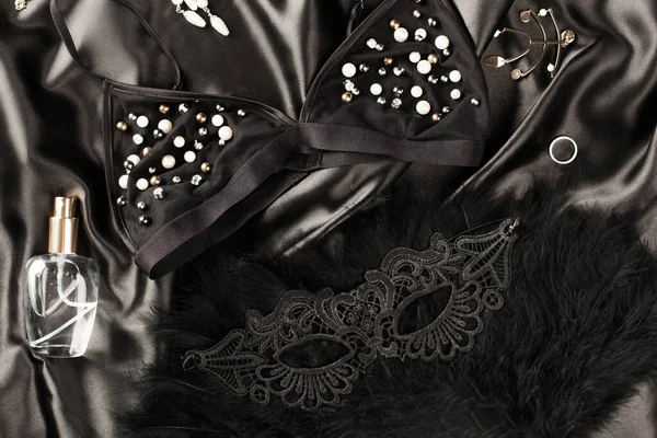 Vista superior de sutiã, perfume e máscara sexual em fundo de cetim preto — Fotografia de Stock