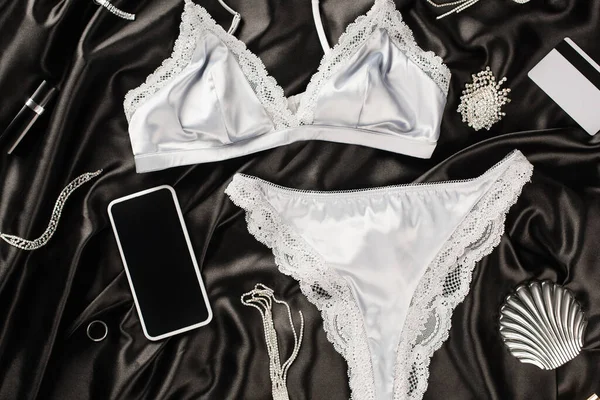 Vista superior de lingerie perto de celular, acessórios e cartão de crédito em fundo de cetim preto — Fotografia de Stock