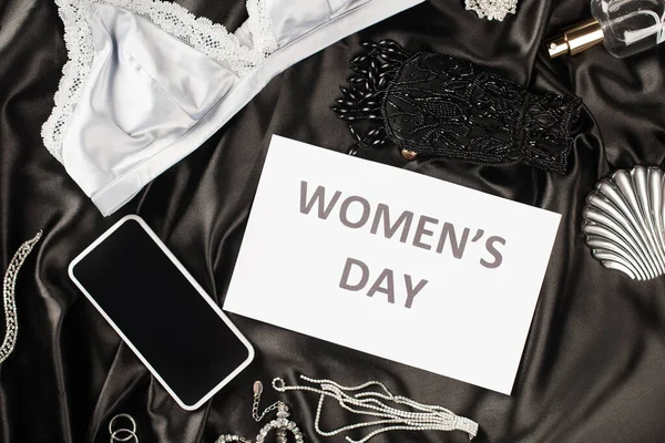 Top view of card with womens day lettering near bra, smartphone and perfume on black satin background — Stock Photo