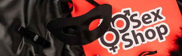 Top view of sexual mask near lipsticks and shopping bag with sex shop lettering on black satin background, banner — Stock Photo