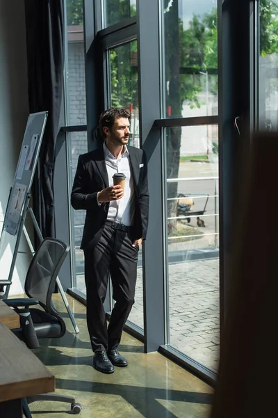 Visão de comprimento total de empresário em terno elegante de pé com café para ir e mão no bolso perto de janelas de escritório — Fotografia de Stock