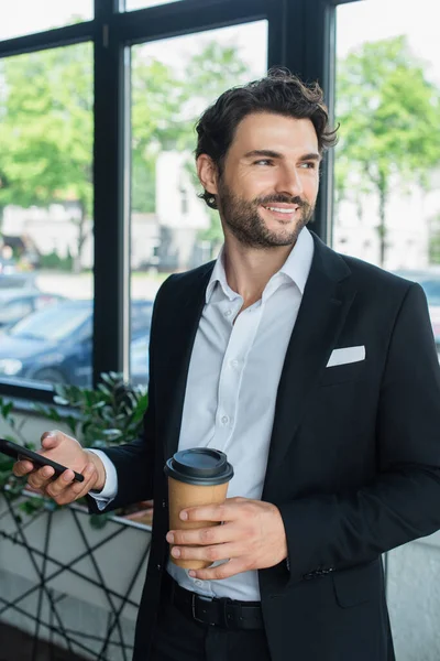 Smiling businessman in black blazer holding smartphone and takeaway drink while looking away in office — Stock Photo