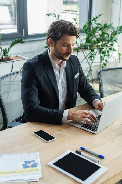 Homem de negócios em elegante formal desgaste digitação no laptop perto de dispositivos na mesa — Fotografia de Stock