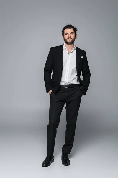 Full length view of man in black elegant suit standing with hands in pockets while looking at camera on grey — Stock Photo