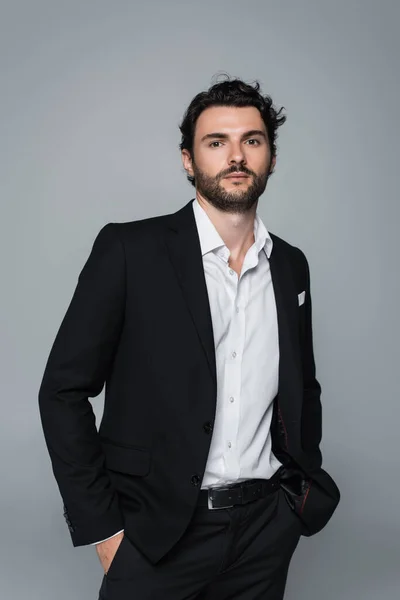 Elegante uomo in blazer nero e camicia bianca in piedi con le mani in tasche isolate sul grigio — Foto stock