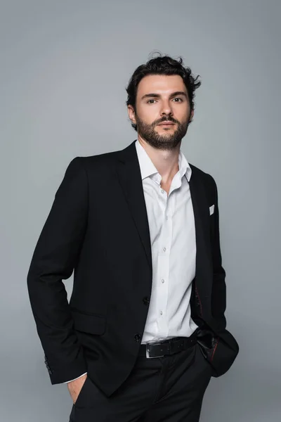 Homme élégant en blazer noir et chemise blanche debout avec les mains dans des poches isolées sur gris — Photo de stock