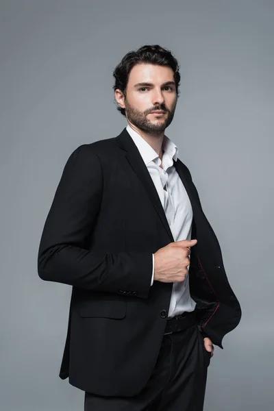 Brunette man in black blazer and white shirt holding hand in pocket isolated on grey — Stock Photo