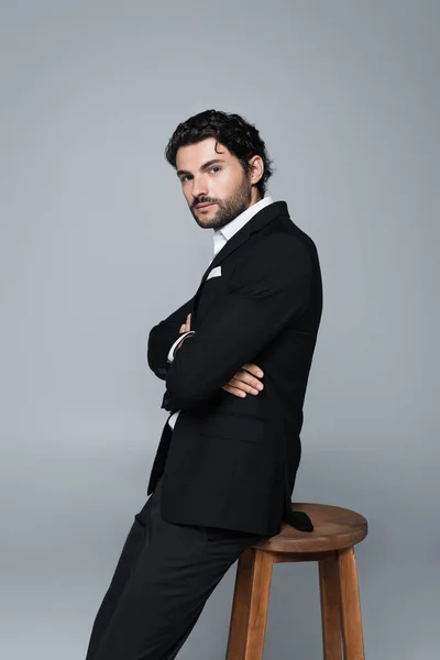 Stylish man in black suit standing near high stool with crossed arms and looking at camera isolated on grey — Stock Photo