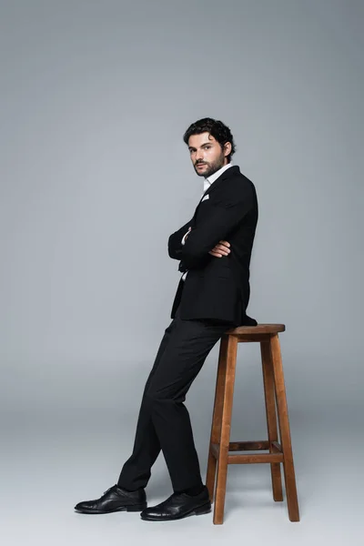 Vue pleine longueur de l'homme élégant en costume noir posant avec les bras croisés près de tabouret haut sur gris — Photo de stock