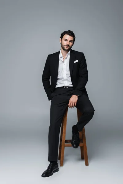 Ganzkörperansicht eines Mannes im schwarzen Anzug, der auf einem hohen Hocker sitzt und in die Kamera auf grau blickt — Stockfoto