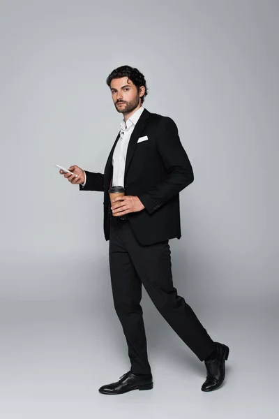 Full length view of businessman in black suit standing with smartphone and paper cup on grey — Stock Photo