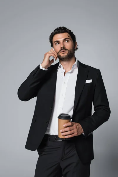 Брюнетка людина в чорному блістері тримає каву, щоб піти і розмовляти на смартфоні ізольовано на сірому — стокове фото