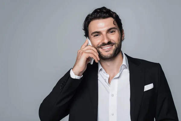 Allegro uomo in blazer nero e camicia bianca che parla su smartphone e guarda la fotocamera isolata su grigio — Foto stock