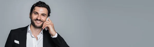 Brunette man in formal wear smiling at camera while talking on mobile phone isolated on grey, banner — Stock Photo