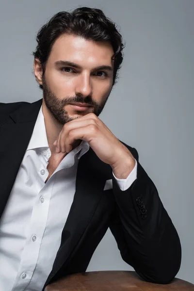 Brunette man in black blazer and white shirt holding hand near face and looking at camera isolated on grey — Stock Photo