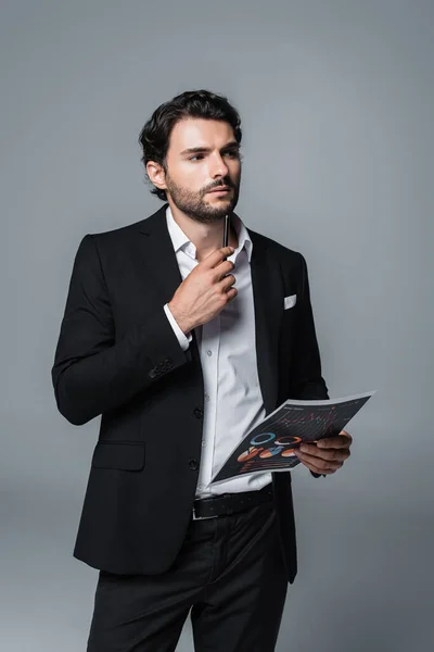 Pensativo hombre de negocios en traje negro con infografías y bolígrafo mientras mira hacia otro lado aislado en gris - foto de stock