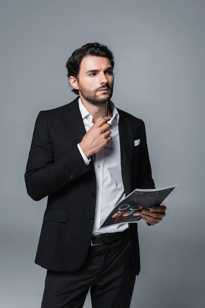 Thoughtful businessman in black suit holding infographics and pen while looking away isolated on grey — Stock Photo