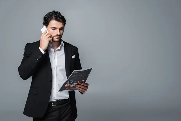 Businessman in formal wear looking at charts and talking on mobile phone isolated on grey — Stock Photo