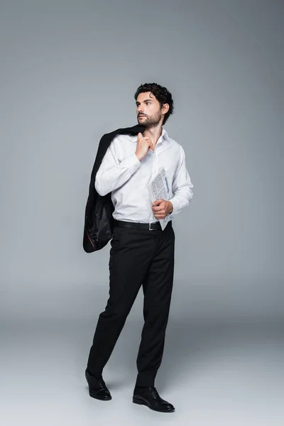 Full length view of businessman holding blazer and newspaper while looking away on grey — Stock Photo