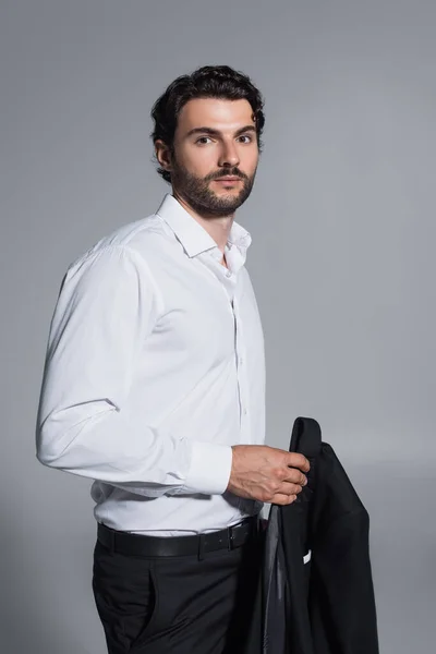 Brunette man in white shirt holding black blazer while looking at camera isolated on grey — Stock Photo