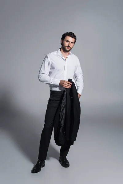 Full length view of man holding black blazer and looking at camera while standing on grey — Stock Photo