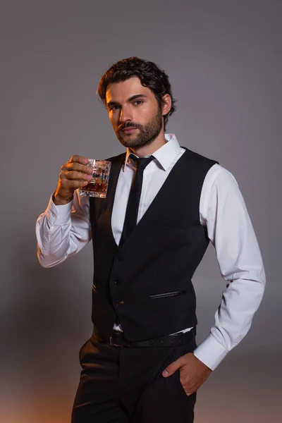 Eleganter brünetter Mann mit einem Glas Whisky im Stehen mit der Hand in der Tasche auf grauem Hintergrund — Stockfoto