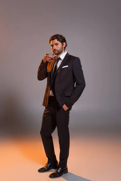 Vista completa del hombre en traje elegante sosteniendo vaso de whisky sobre fondo gris con luz - foto de stock