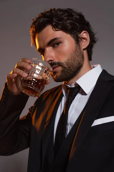 Stylischer Mann im schwarzen Anzug trinkt Whisky, während er in die Kamera schaut, isoliert auf grau — Stockfoto