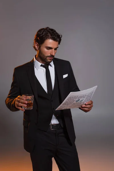 Uomo in abito elegante nero che legge giornale mentre tiene un bicchiere di whisky su sfondo grigio — Foto stock